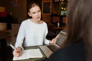 A woman with a laptop and a notebook talking to a another woman 